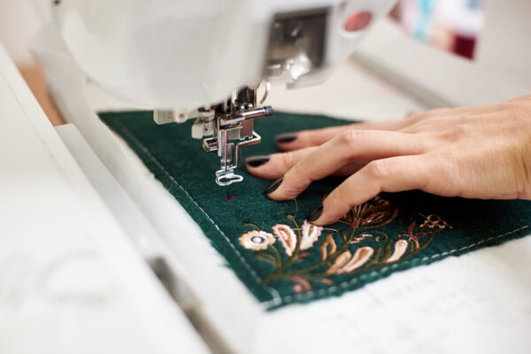 Close-up of female manicured hand working on sewing machine creating colorful floral pattern on dark green textile detail. Modern technology, fashion and traditional clothing manufacturing concept.
